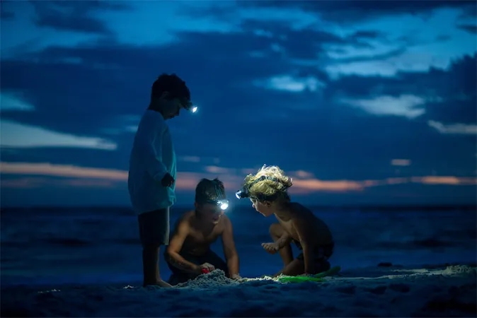 Trois enfants jouent avec une lampe de poche sur la plage, riant et s'amusant sous le ciel étoilé.