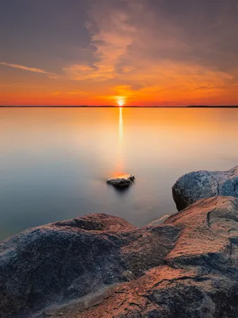 Coucher de soleil sur l'eau, avec des rochers au premier plan, créant une scène paisible et colorée.